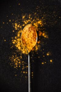 From above of creative composition of bright ground turmeric on metal tablespoon spilled on black table